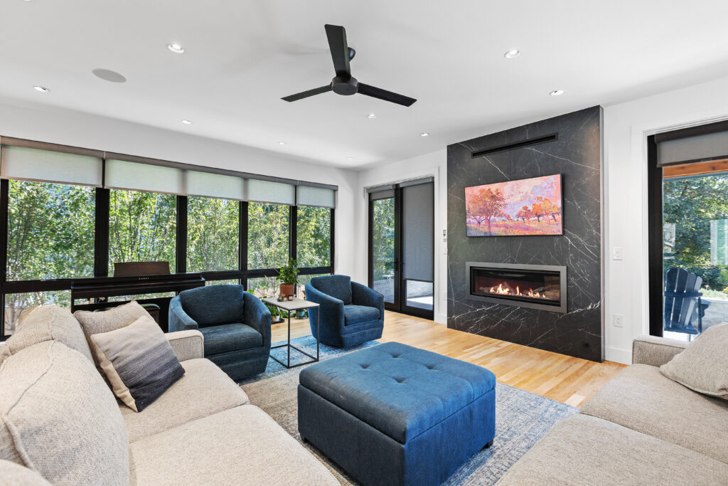 A living room with a black marble electric fireplace with a fire in the grate, dark blue fabric chairs and ottoman, cream sofa, and a wall of bright windows.