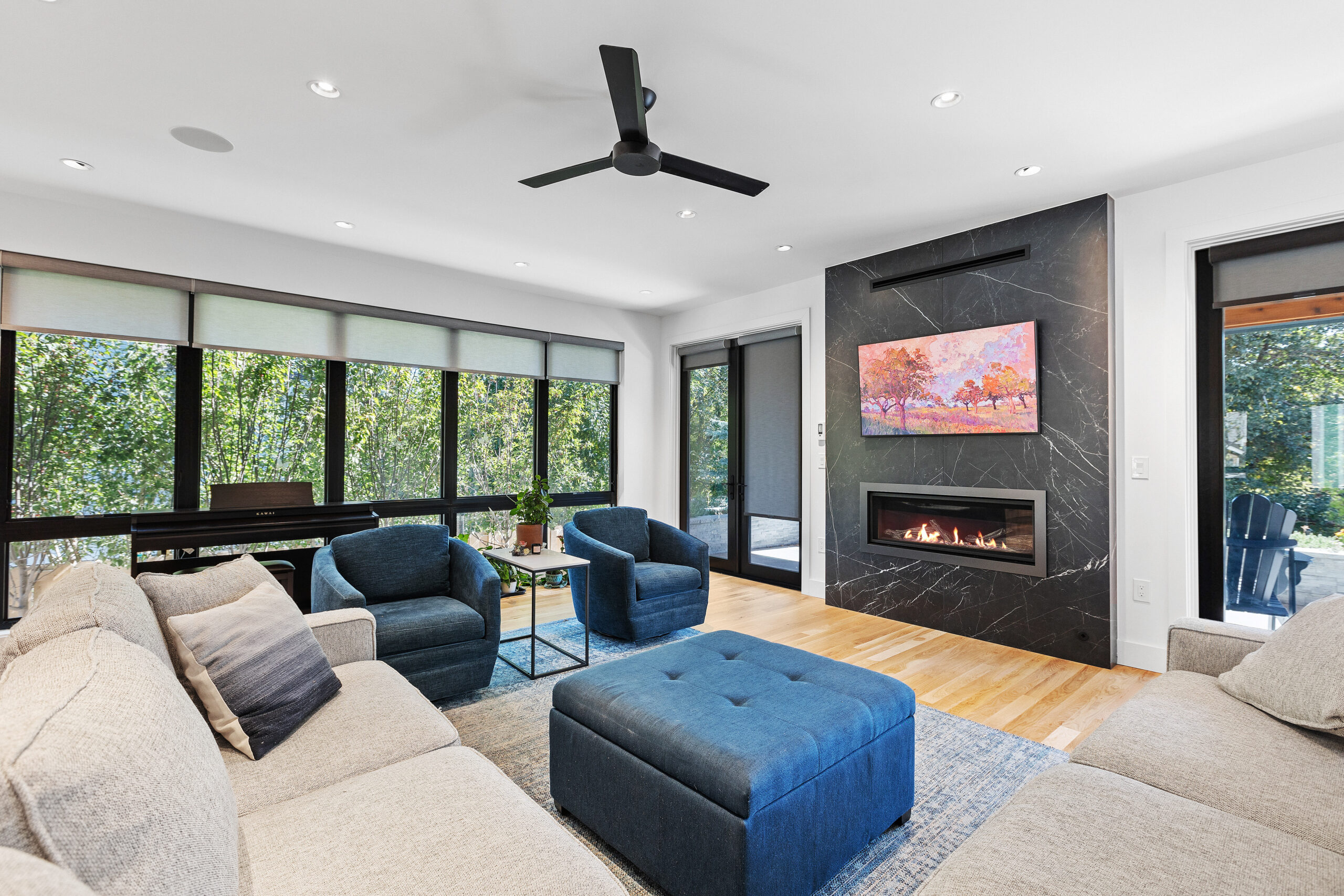 A living room with a black marble electric fireplace with a fire in the grate, dark blue fabric chairs and ottoman, cream sofa, and a wall of bright windows.
