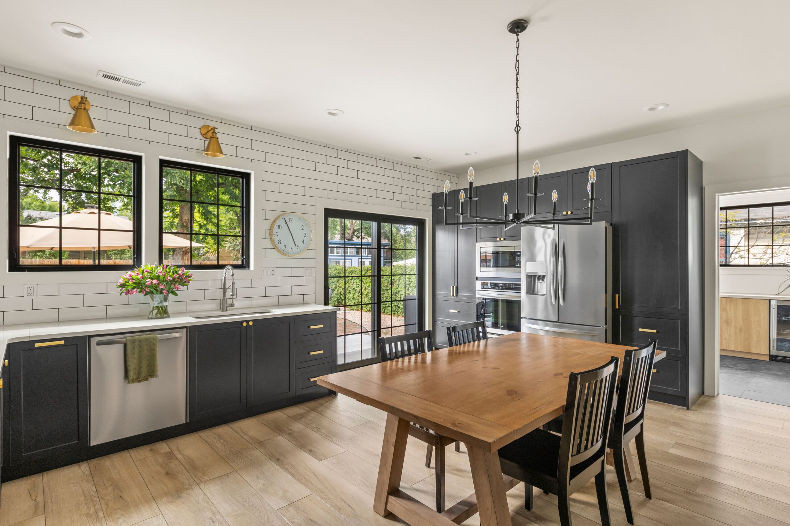 A large indoor kitchen with white tiled walls, wood floor, wood table, dark gray cabinets, large windows, and pink flowers on a marble countertop.