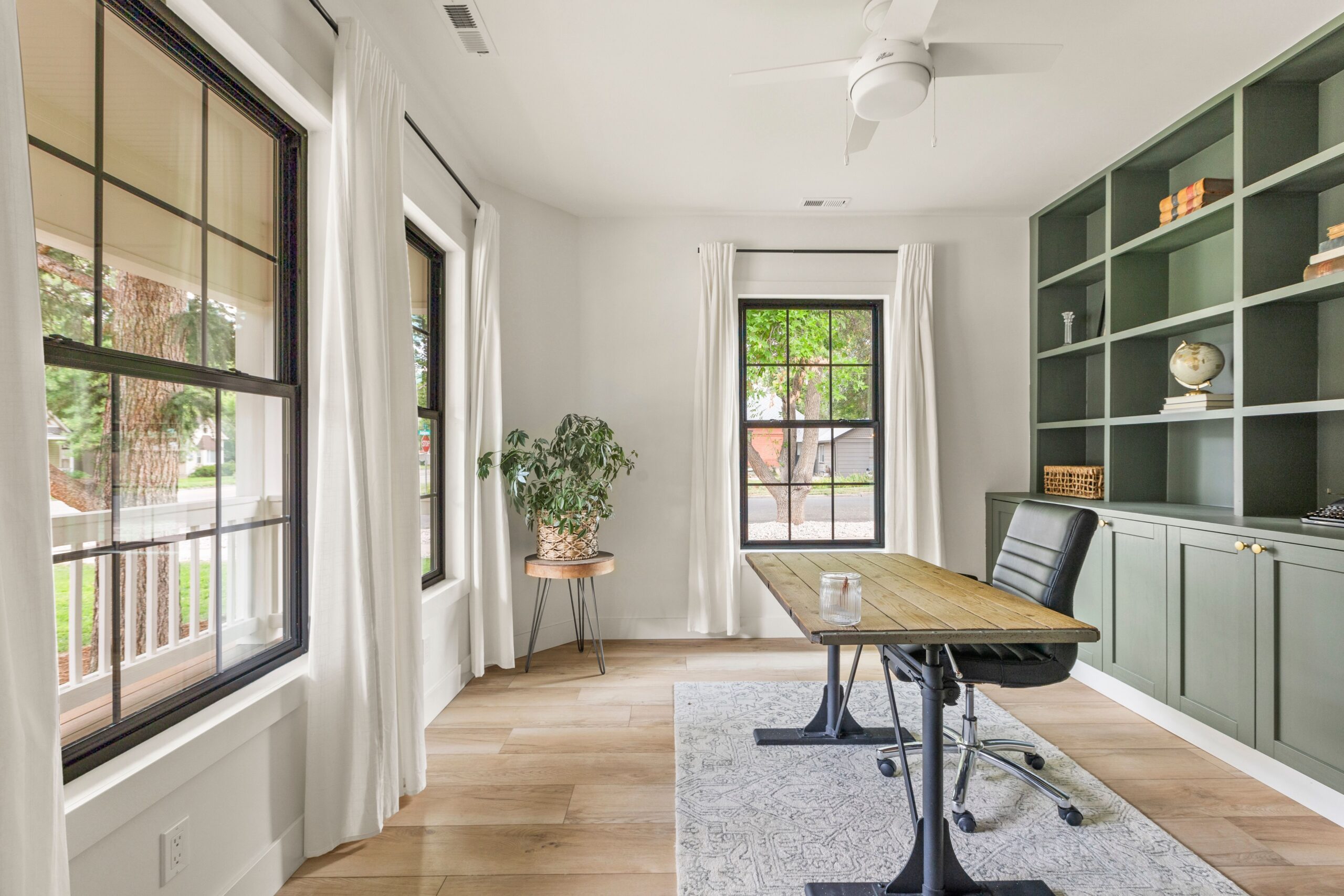 An office in a house with large windows, wood floors, a desk, and an olive green bookshelf.
