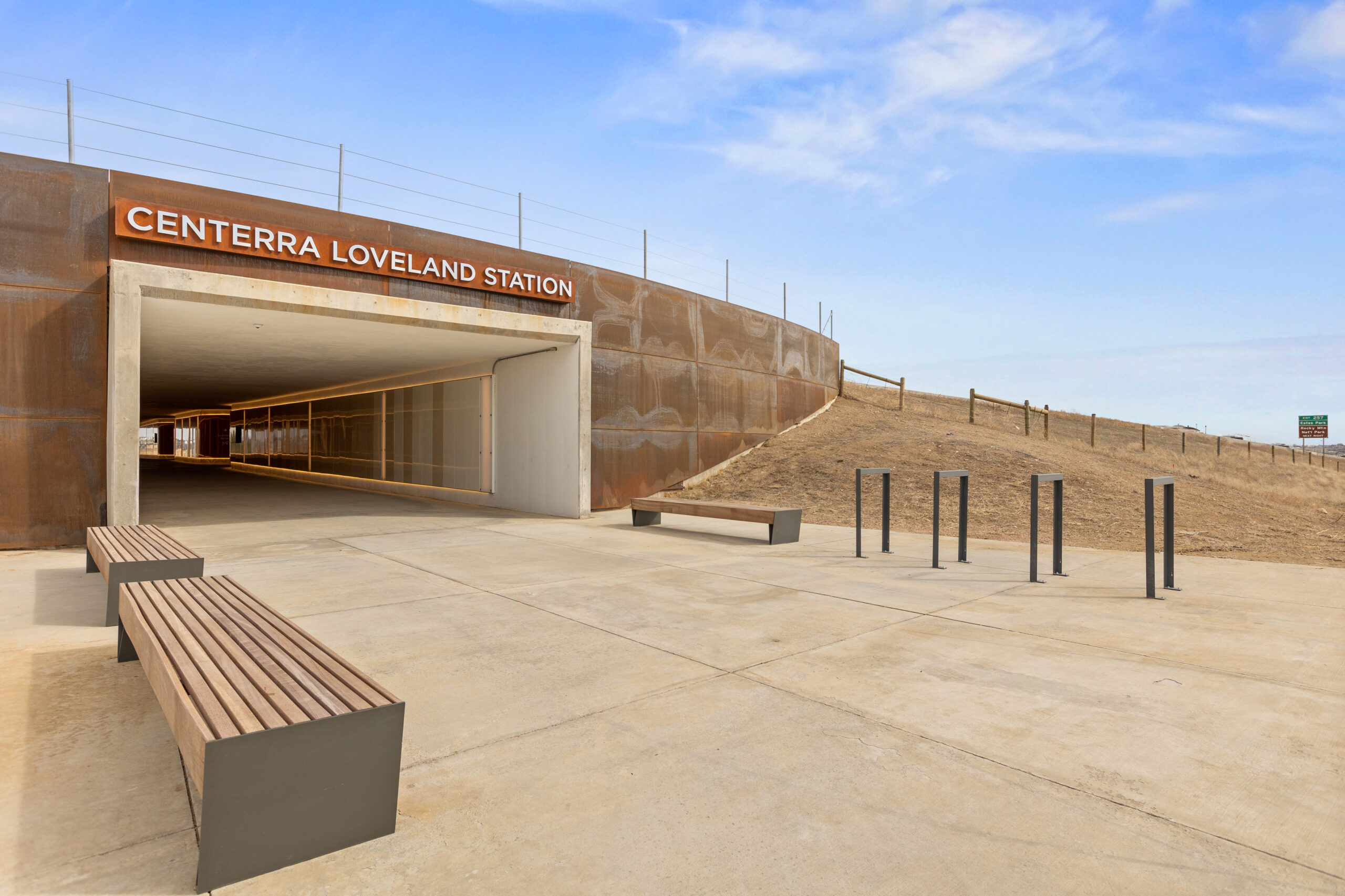 A tunnel from the outside with blue sky above, wooden benches, bike racks, and a sign "Centerra Loveland Station".